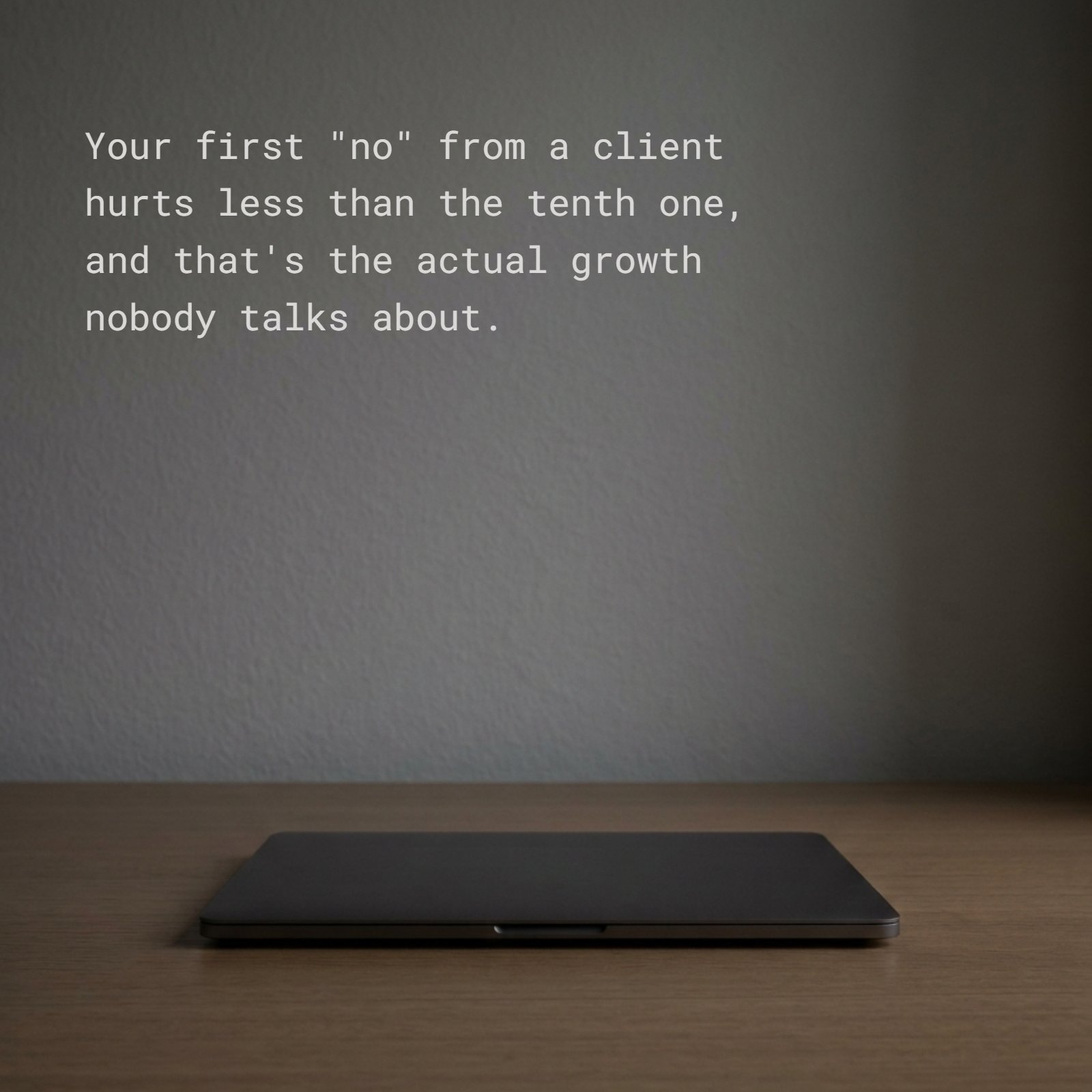 A minimalist photo of a single closed dark grey laptop resting on a plain wooden desk in dim evening light. Superimposed on the clean negative space above the laptop is the text quote: Your first "no" from a client hurts less than the tenth one, and that's the actual growth nobody talks about.