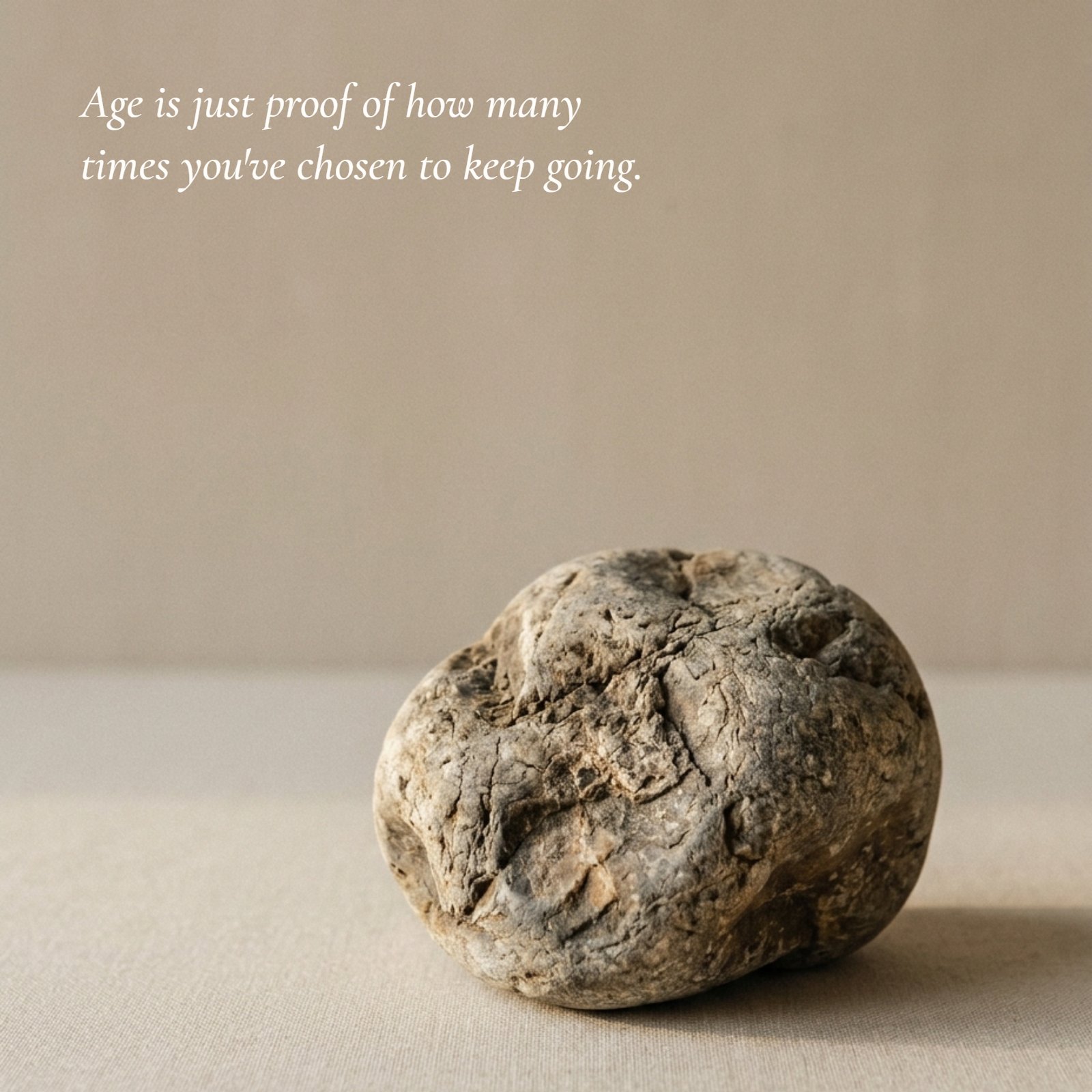 Minimalist photo of a single weathered stone with deep textures on a plain neutral surface. Superimposed is the text of the quote: 'Age is just proof of how many times you've chosen to keep going.' in a classic serif font style.
