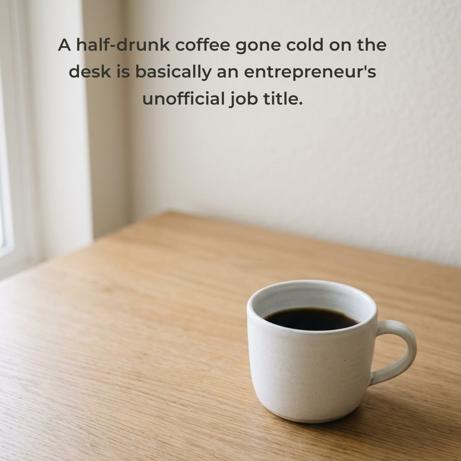 A minimalist photo showing a white ceramic coffee cup sitting on a clean wooden desk with the text overlay 'A half-drunk coffee gone cold on the desk is basically an entrepreneur's unofficial job title.'