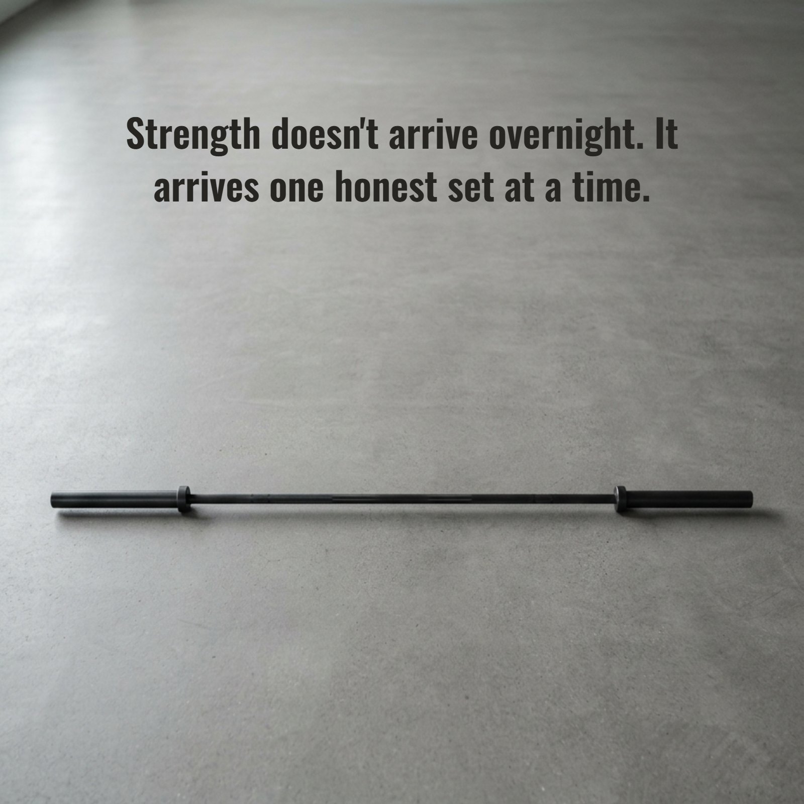 A minimalist photograph of a single black Olympic barbell resting on a plain grey concrete gym floor under soft light, featuring the quote Strength doesn't arrive overnight. It arrives one honest set at a time.