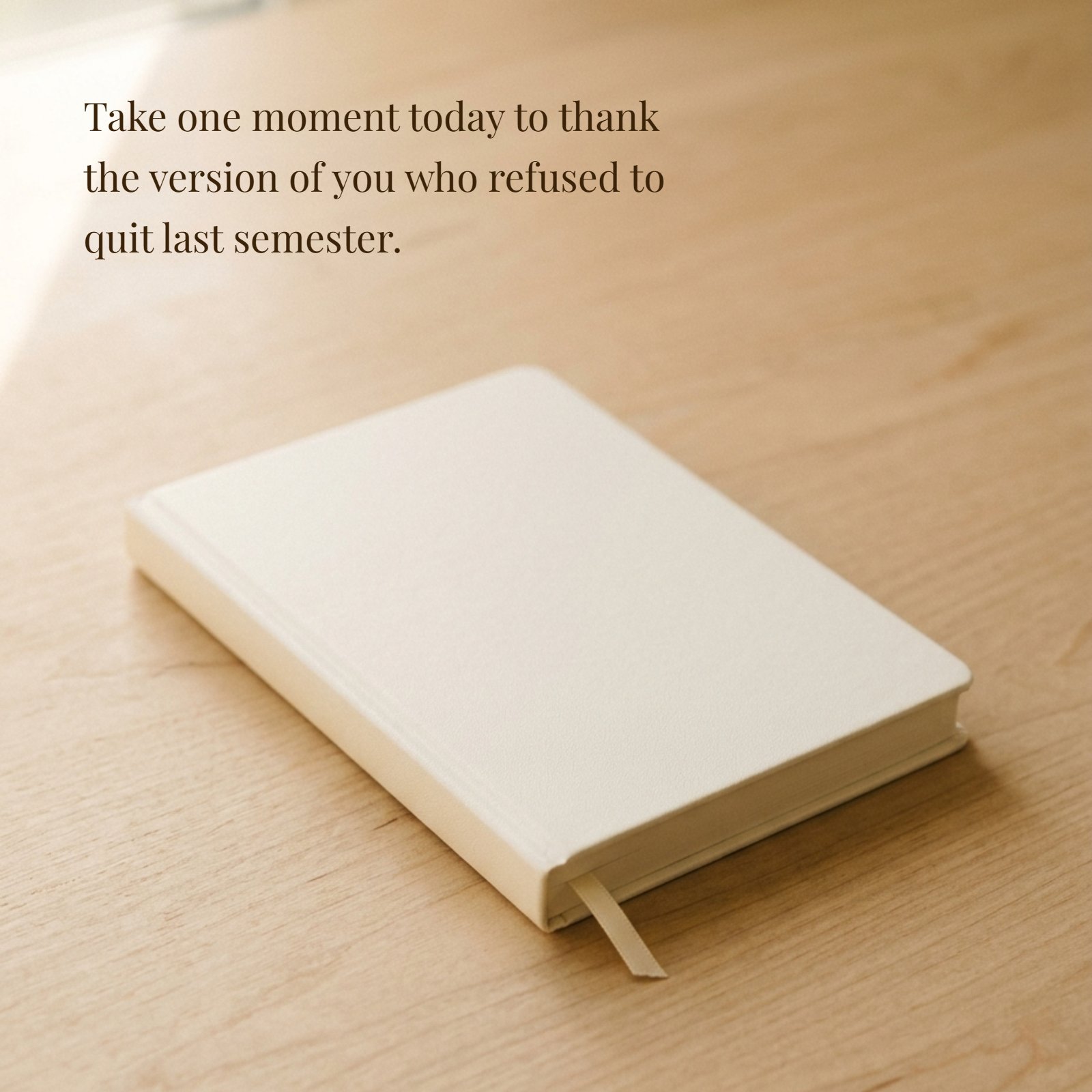 A white closed notebook on a wooden desk featuring the inspirational quote: Take one moment today to thank the version of you who refused to quit last semester.