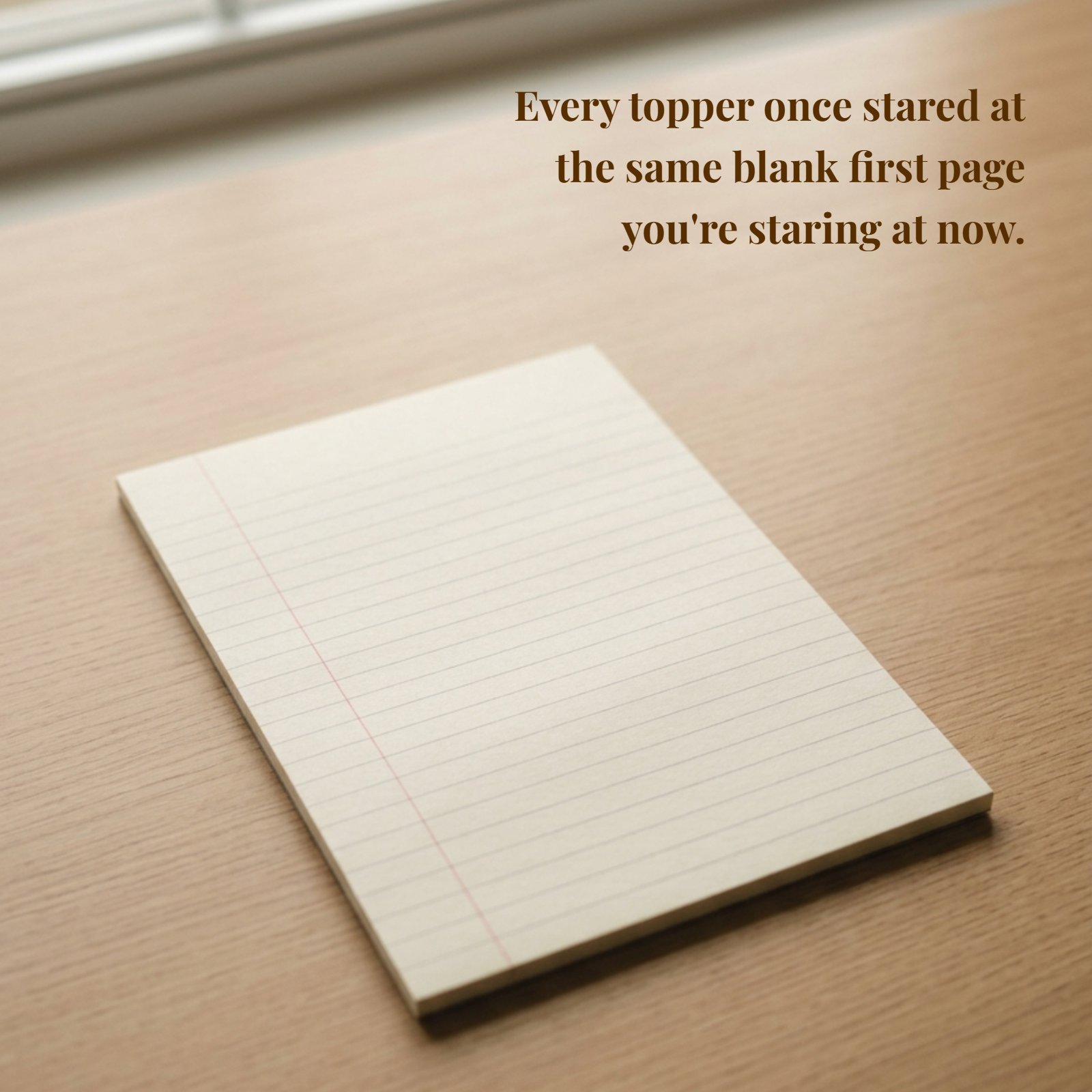 Minimalist photo of a single lined notebook page on a light wooden desk with the quote “Every topper once stared at the same blank first page you're staring at now.”