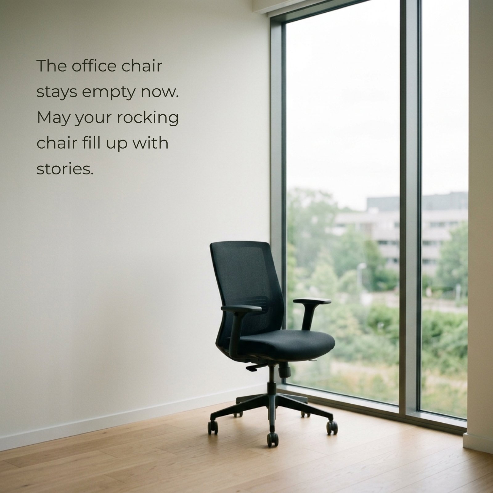 Minimalist 1:1 photograph featuring a modern empty black mesh office chair on a light wood floor next to a tall, floor-to-ceiling window. The quote, “The office chair stays empty now. May your rocking chair fill up with stories,” is written on the large plain wall area to the left. The window shows a soft, neutral, out-of-focus view of green trees and distant buildings.