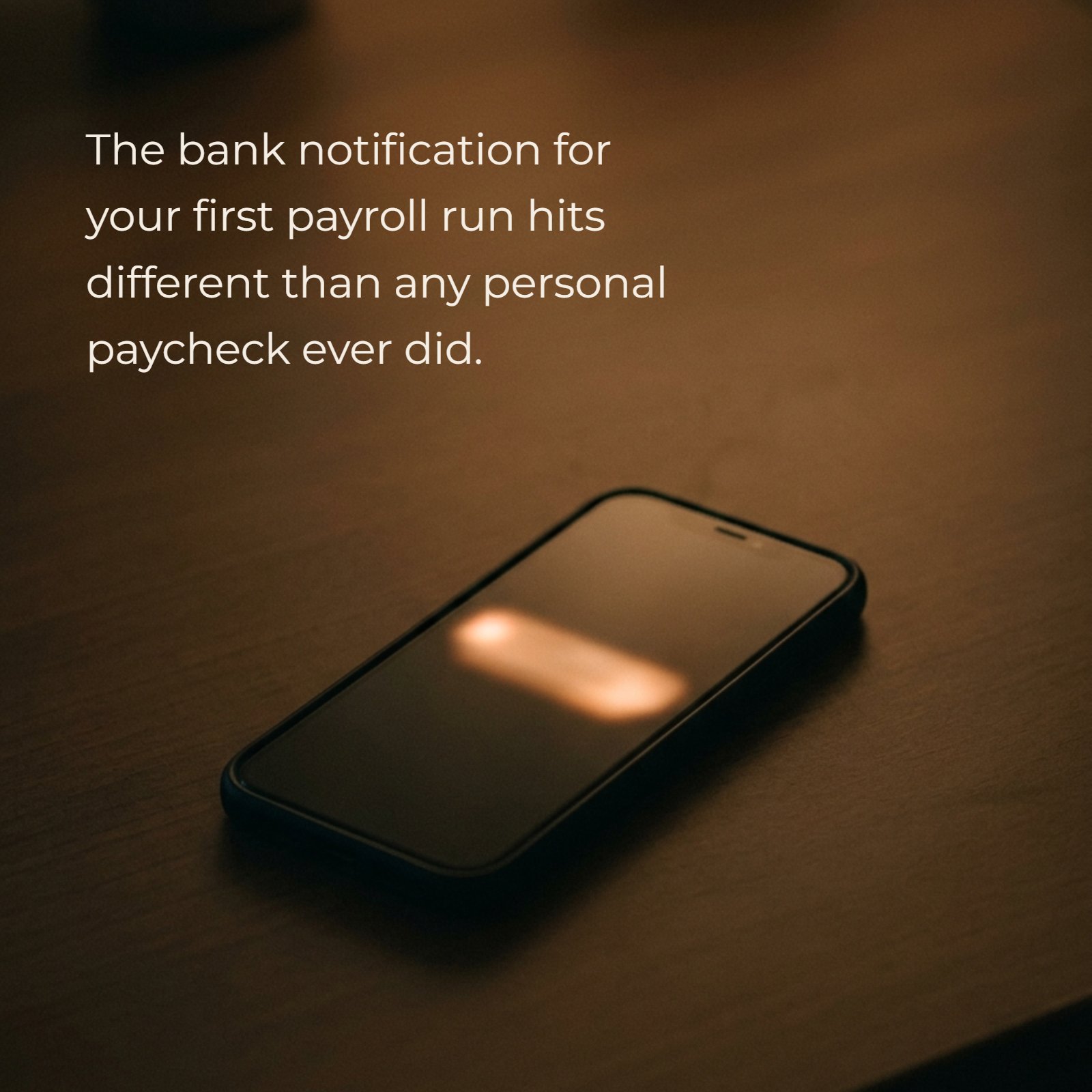 A minimalist photo of a single smartphone lying face-up on a plain desk under warm, dim light. A single, distinct, soft notification glow illuminates a small portion of the screen, indicating a notification without displaying any text. The phrase “The bank notification for your first payroll run hits different than any personal paycheck ever did.” is overlaid in a clean font.