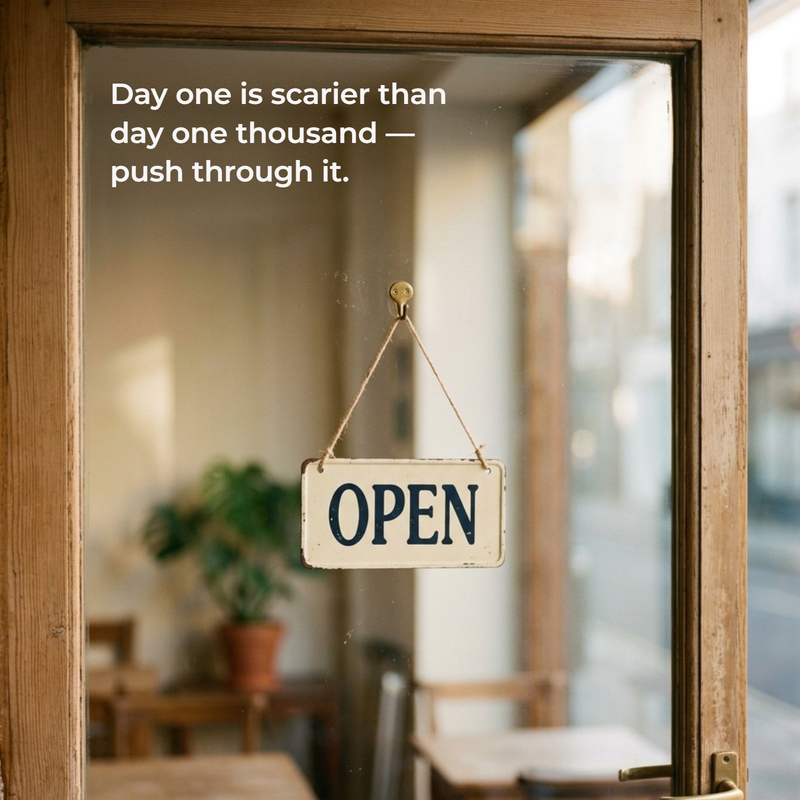 A minimalist photo of a wooden-framed glass door with a classic white "Open" sign hanging. Overlaid text reads, Day one is scarier than day one thousand — push through it.