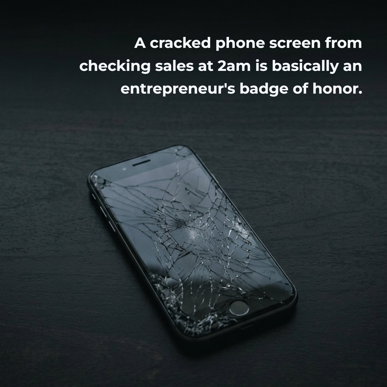 Minimalist 4:5 photograph on a dark background showing a smartphone with a deeply cracked screen lying on a dark desk. The overlay text reads: 'A cracked phone screen from checking sales at 2am is basically an entrepreneur's badge of honor.'