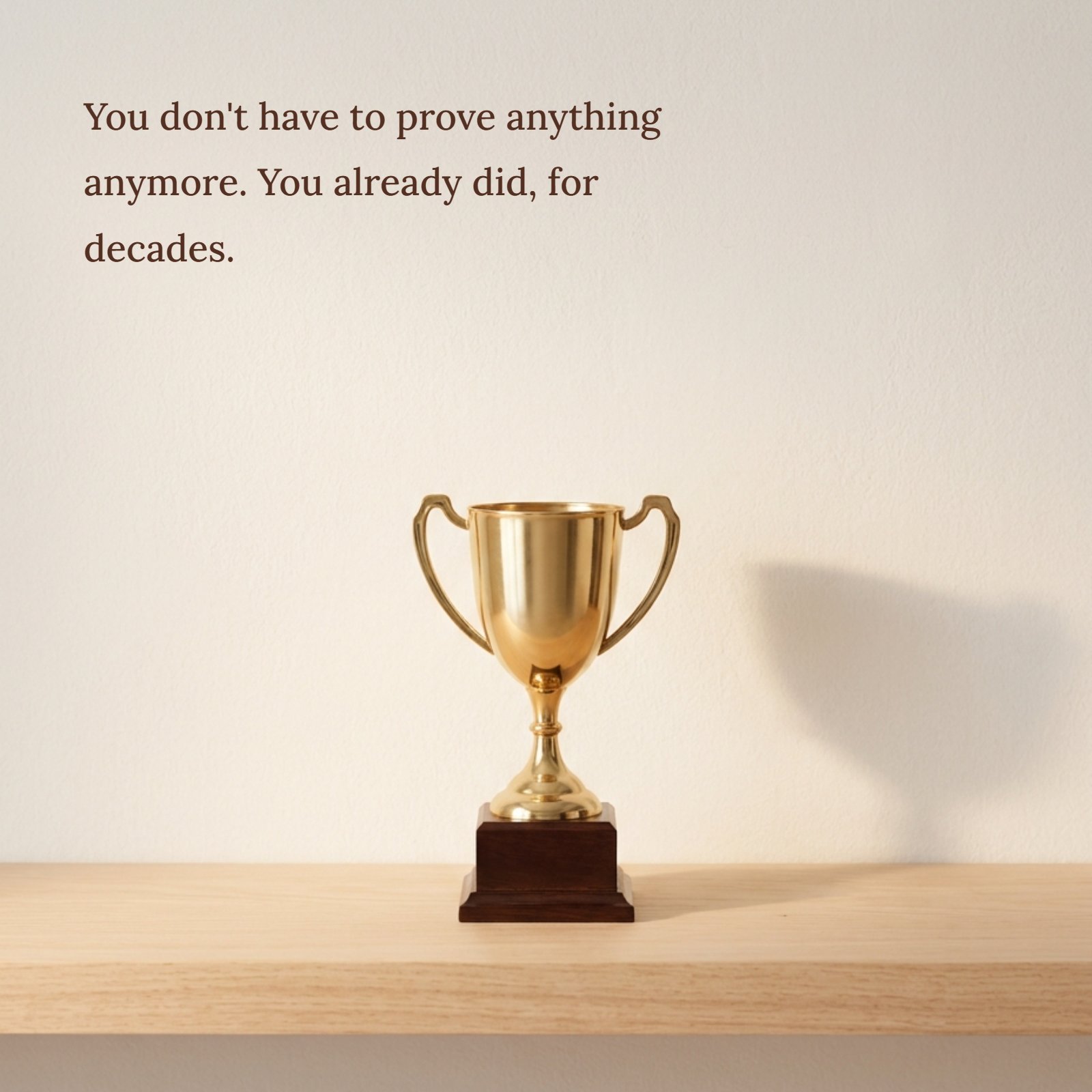 A clean, well-lit photograph features a classic golden trophy cup resting on a simple wooden shelf against a textured light-colored wall. Above the trophy, the motivational quote is clearly visible in an elegant dark font: 'You don't have to to prove anything anymore. You already did, for decades.' The scene is simple and focuses on the idea of long-term achievement and validation.