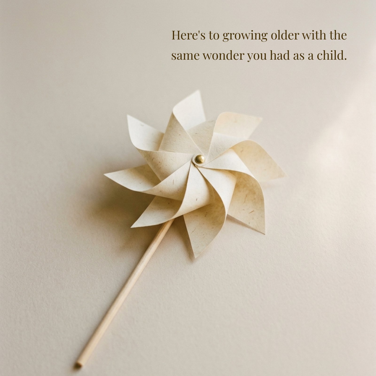 A minimalist photo of a cream-colored paper pinwheel on a plain beige surface with soft natural light, perfect for featuring the quote: 'Here's to growing older with the same wonder you had as a child.'