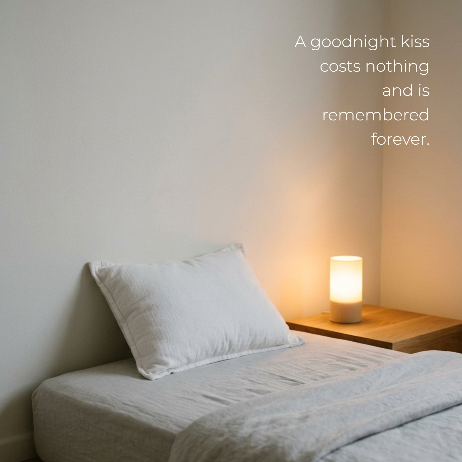 Minimalist photo of a soft white pillow on a bed with a warm glowing lamp, featuring the text overlay: A goodnight kiss costs nothing and is remembered forever.