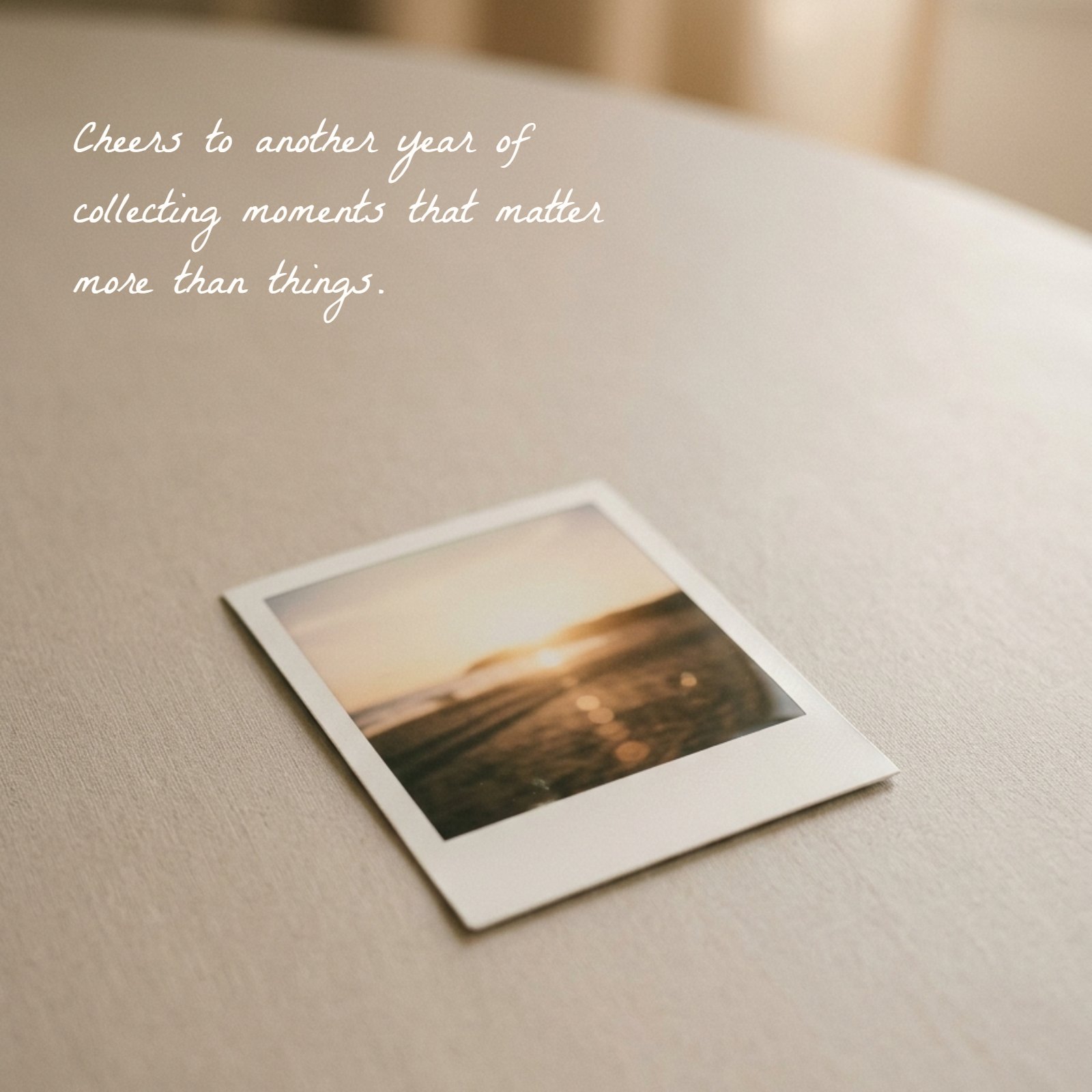 A minimalist photo of a single polaroid-style picture with a sunset scene resting on a light neutral table surface. Text overlay reads "Cheers to another year of collecting moments that matter more than things."