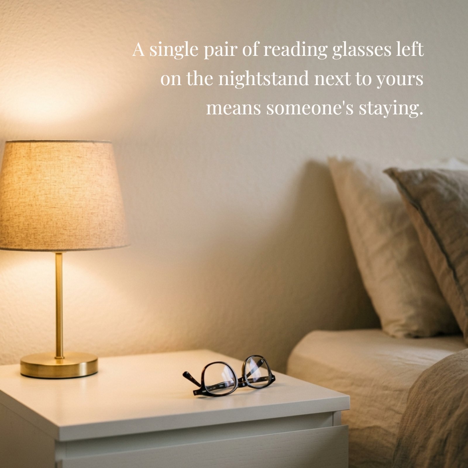A minimalist photo showing a single pair of reading glasses resting on a nightstand illuminated by soft, warm light from a lamp. The minimalist design emphasizes the message that these glasses mean someone is staying.