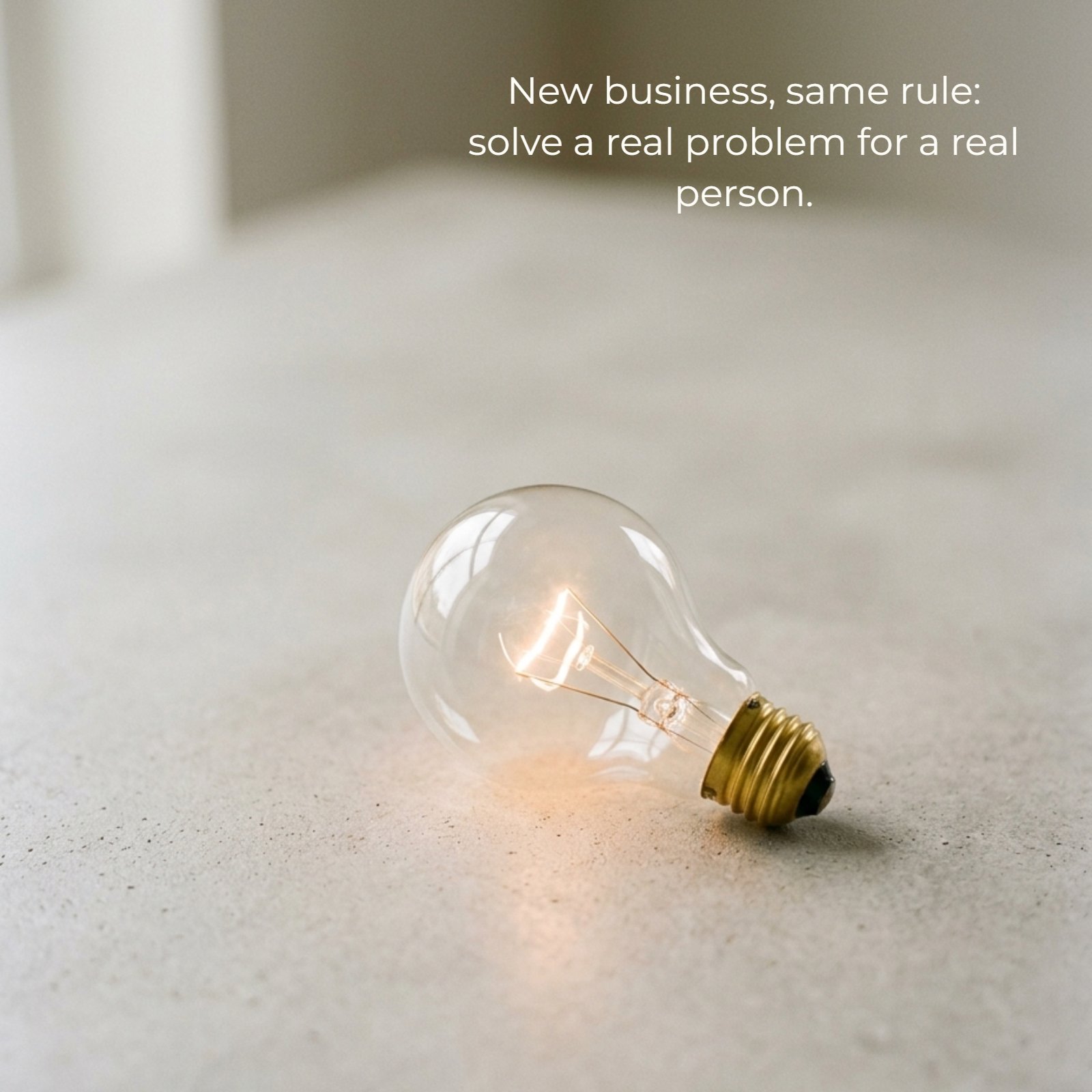 Minimalist photo of a single glowing lightbulb on a plain neutral desk, representing a new idea. It features the text: New business, same rule: solve a real problem for a real person.