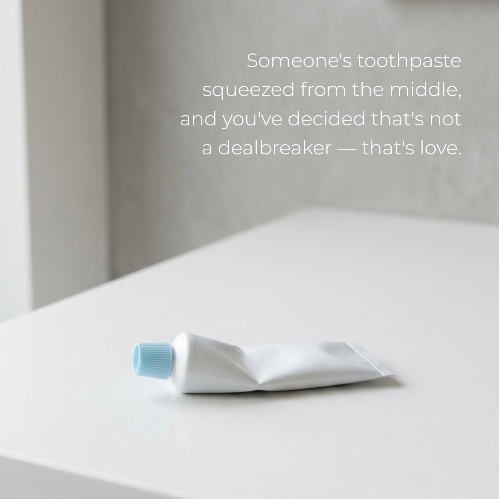 A minimalist 1:1 photograph featuring a single, blank white toothpaste tube on a plain bathroom counter, squeezed noticeably from the middle. Soft natural light illuminates the scene with abundant clean negative space. Overlaid text reads: ‘Someone's toothpaste squeezed from the middle, and you've decided that's not a dealbreaker — that's love.’