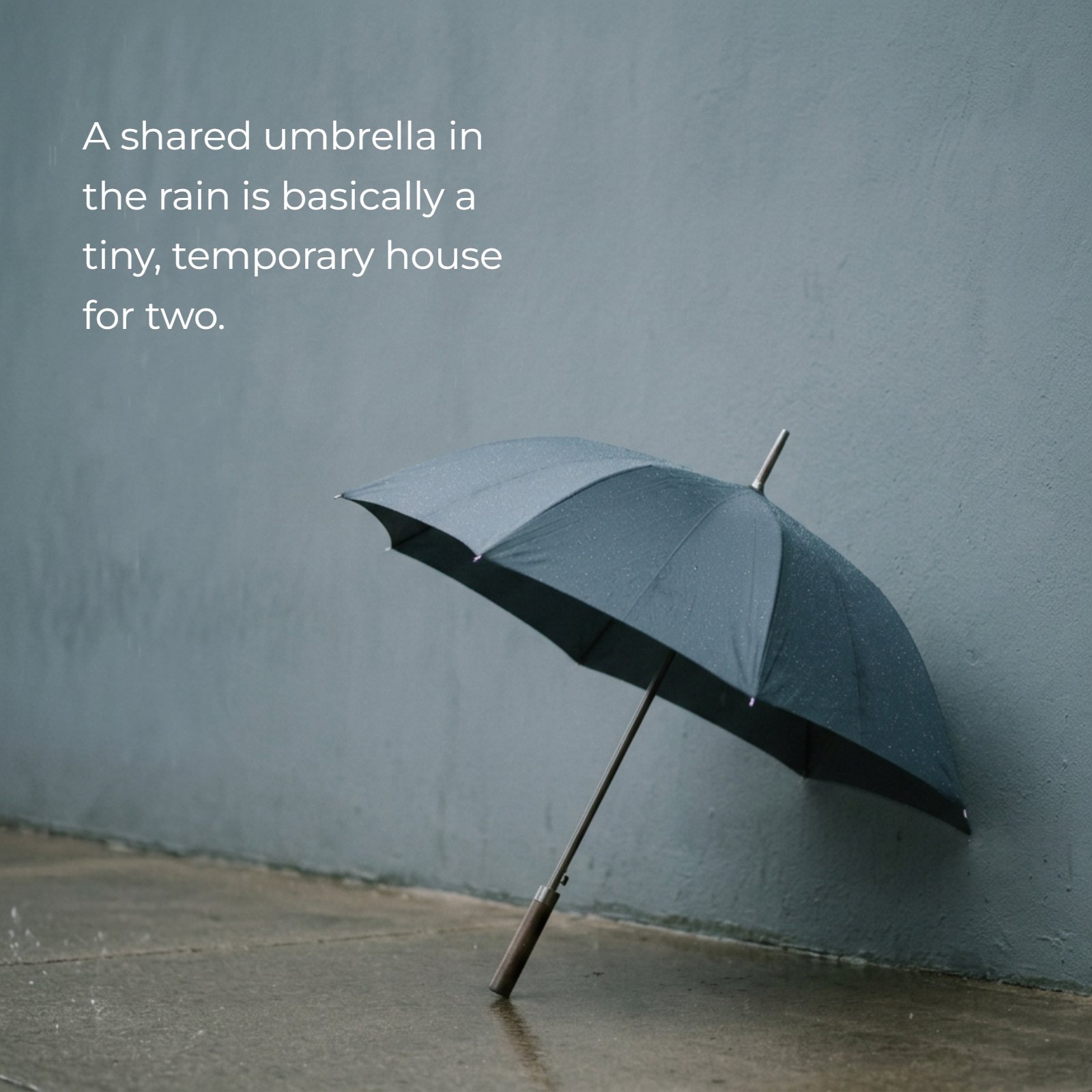 A minimalist photo of a wet, dark umbrella leaning against a grey-blue wall with the text overlay: "A shared umbrella in the rain is basically a tiny, temporary house for two."