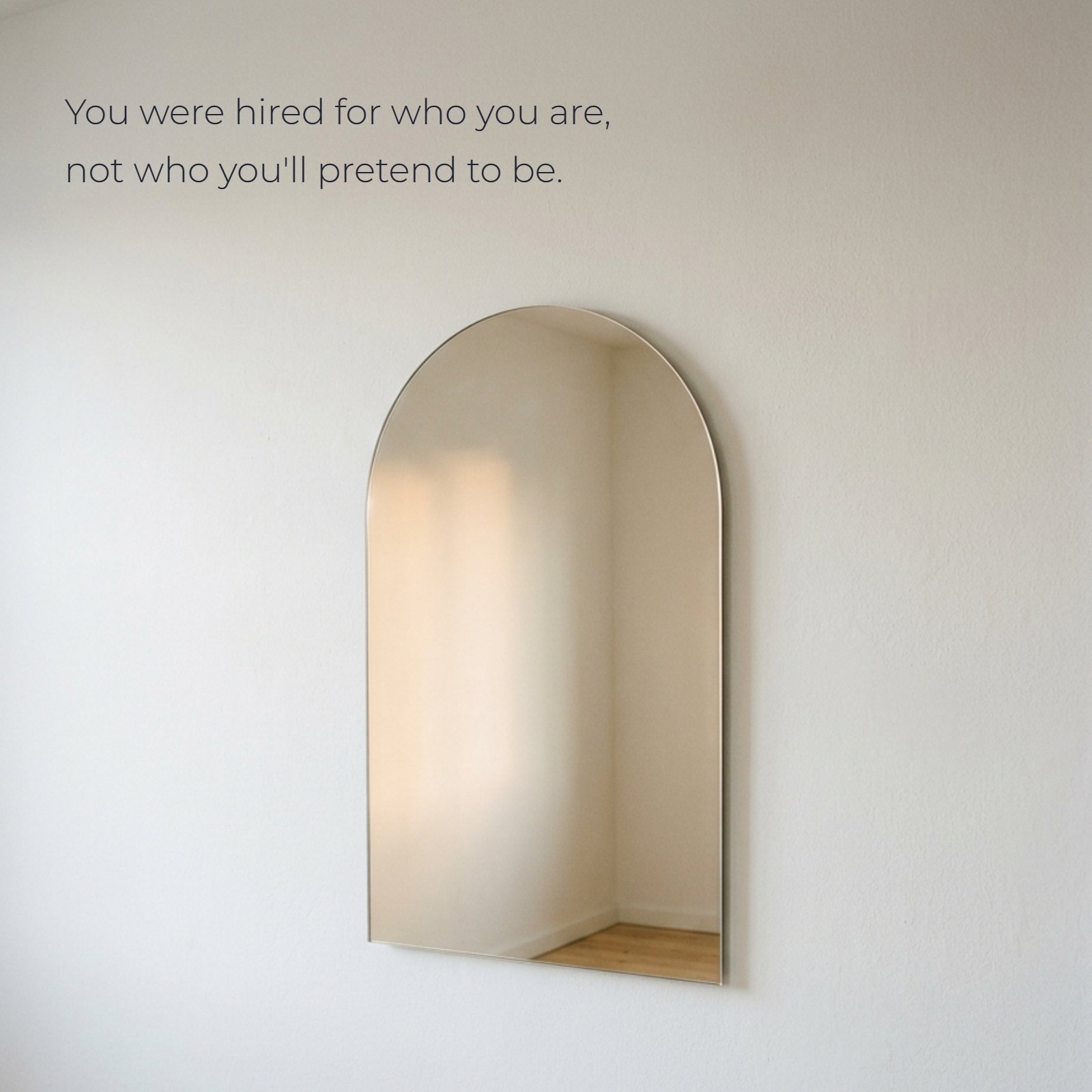 Minimalist 1:1 photograph of an arched mirror on a white wall, reflecting soft, warm light and clean negative space. Text overlay reads: You were hired for who you are, not who you'll pretend to be.