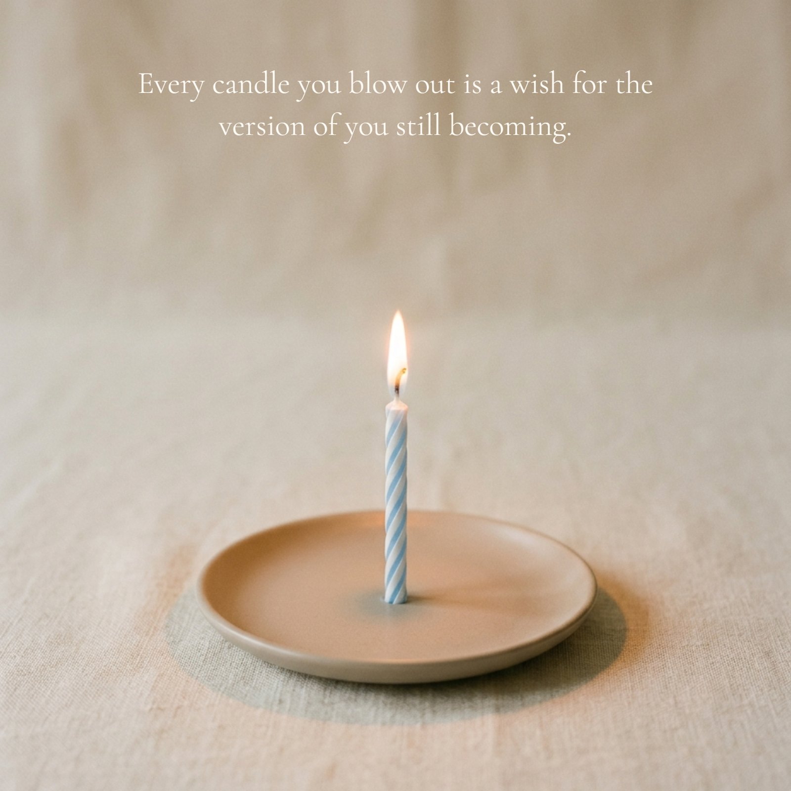 A single, slender, lit blue-and-white striped birthday candle on a small terracotta dish resting on a neutral linen surface. The soft, warm glow of the candle highlights the textured background, with clean negative space around it. Text is overlaid on the image, reading: "Every candle you blow out is a wish for the version of you still becoming."