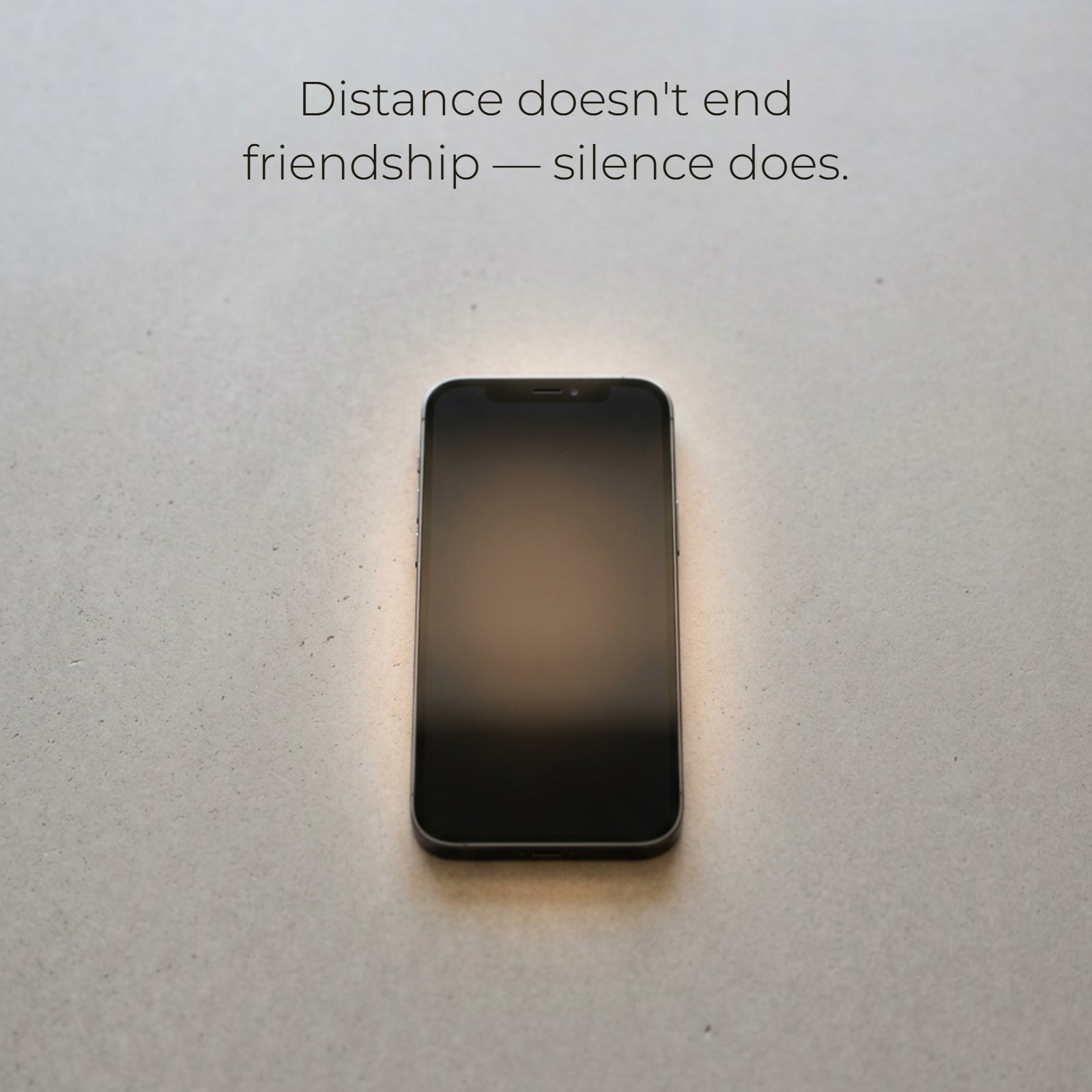 A photograph featuring a black smartphone lying face-up on a grey textured concrete surface, subtly backlit with a soft, warm glow. The minimalist composition includes the quote: 'Distance doesn't end friendship — silence does' in elegant white text.