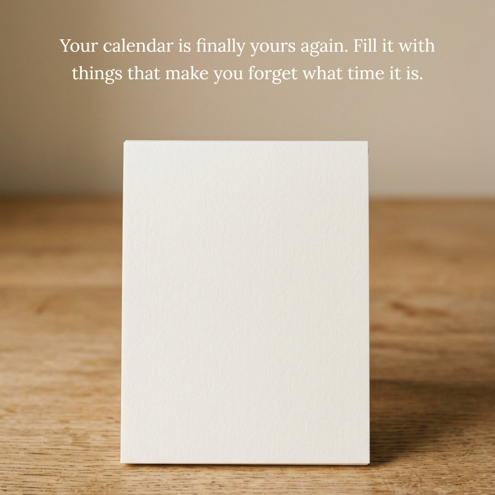 A minimalist photo of a single blank off-white paper calendar page standing on a plain wooden desk under soft warm light. Printed in elegant font on the page is the retirement quote: "Your calendar is finally yours again. Fill it with things that make you forget what time it is."