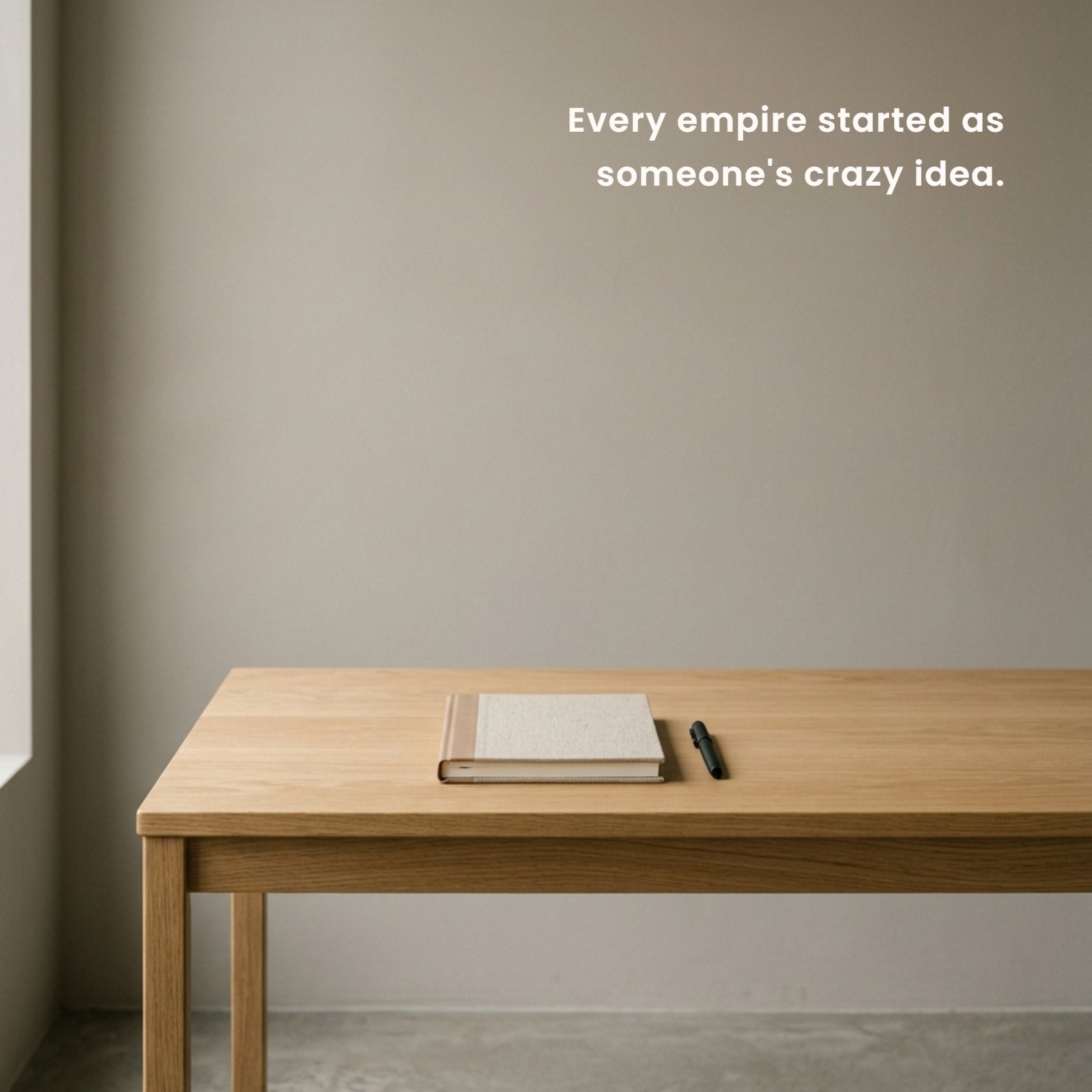 A minimalist 1:1 square photo of a light wood desk against a warm grey wall, containing only a notebook and pen. Overlaid on the spacious grey background is the quote, "Every empire started as someone's 'crazy' idea."