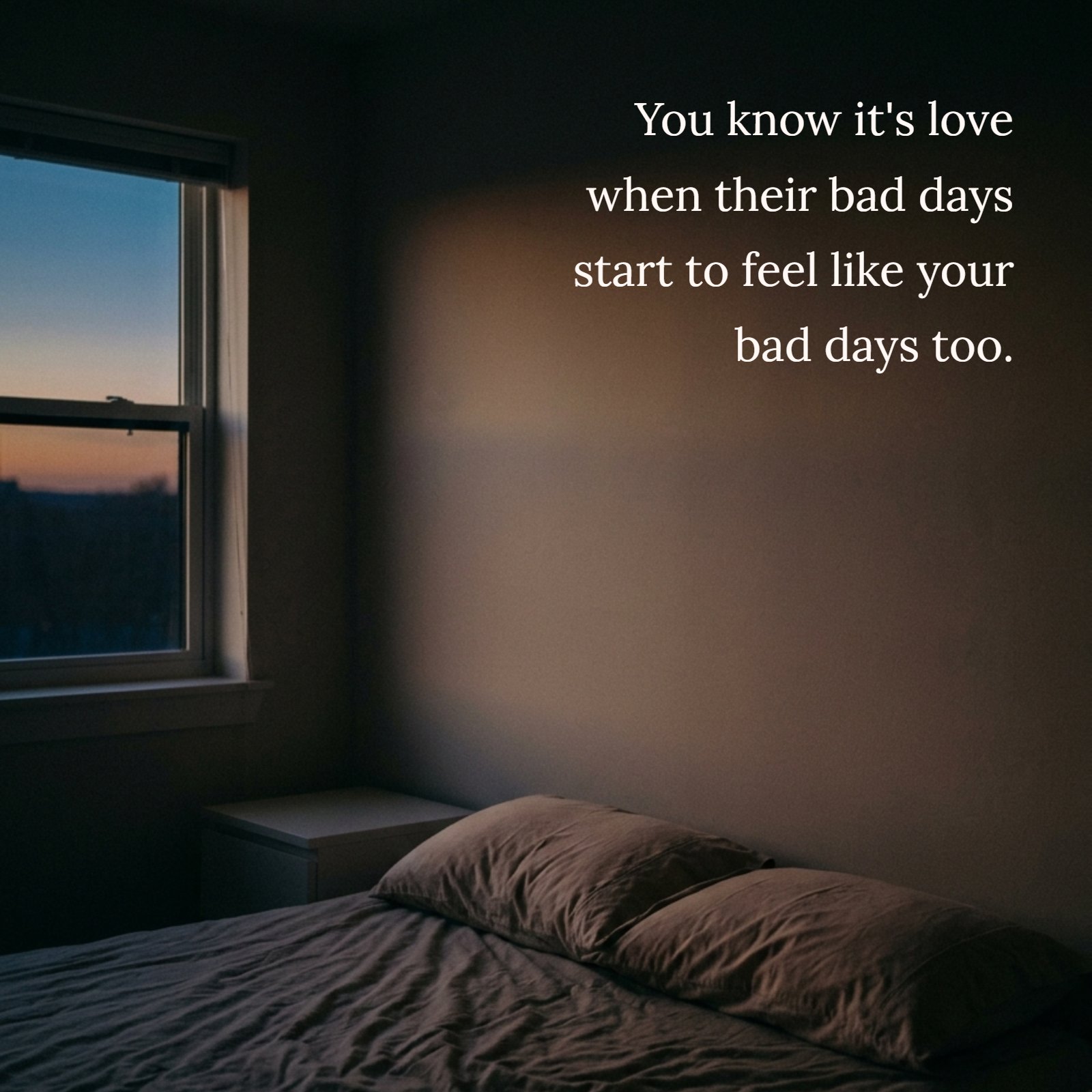 A dim bedroom at dusk features two pillows side-by-side on a bed, with soft twilight filtering through a window. Large shadowed wall space holds the quote, "You know it's love when their bad days start to feel like your bad days too."